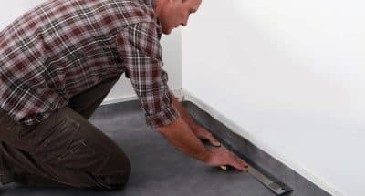 Man in checkered shirt uses long metal tool on the corners of a black linoleum floor.