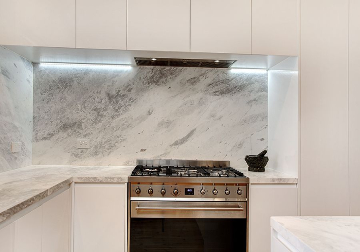 Bright kitchen with white cabinetry, marble benchtop and splashback and stainless steel oven/cooktop.