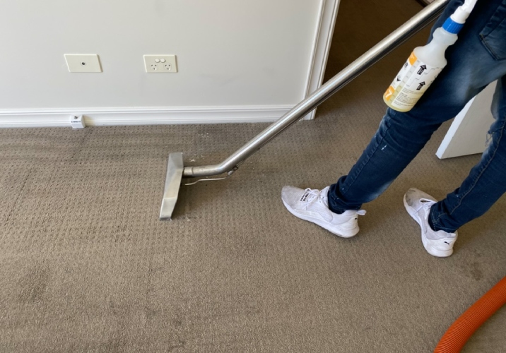 Brown carpet being vacuumed by person in blue jeans and white sneakers.