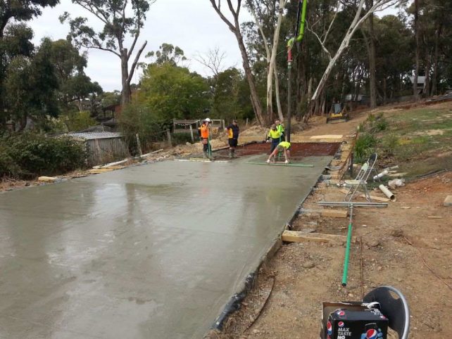 Five professional concreters use a leveller and trowel to smooth out and level concrete for a large driveway in area surrounded by trees.