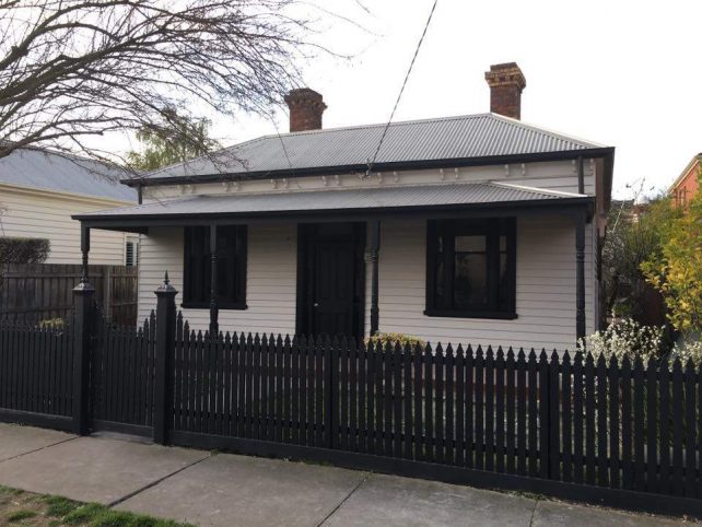 Black picket fence in front of a white weatherboard house with black trims and roof.