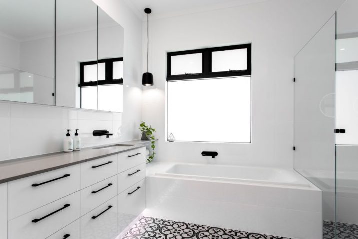 All white tiled bathroom with black fittings and bath centred beneath a window.