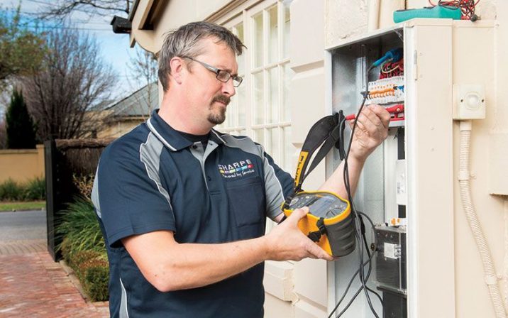 Man with glasses holding device plugged into an electrical box of a white house. 