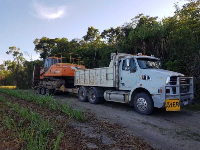 Large white truck with orange excavator on the trailer driving on a dirt road.