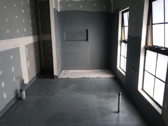 A bathroom construction in its early stages with no fixtures, only plastered walls and dark grey waterproofed floors.