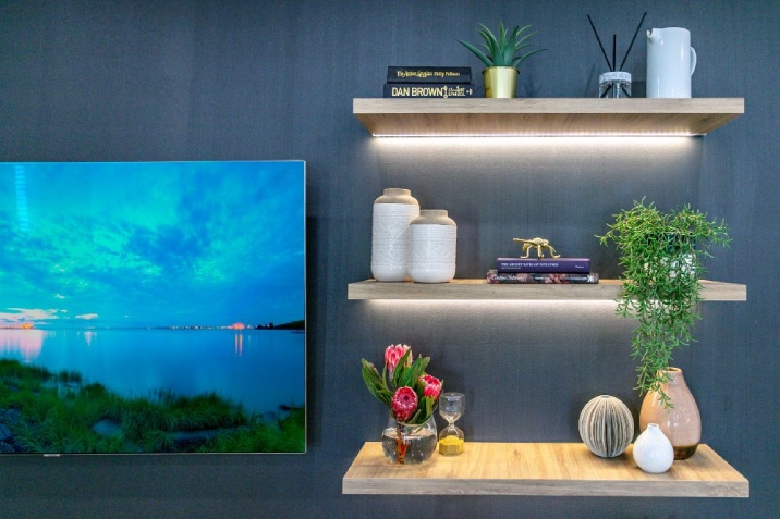 Three neon-lit timber shelves with decorative pieces on each next to a landscape painting on a blue wall.