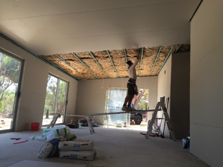 A man uses a ladder and a plank to stand on and push insulation into a ceiling.