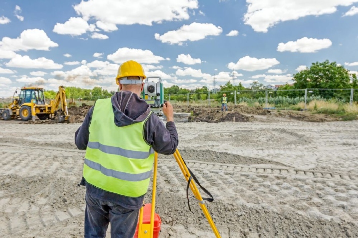 Man in yellow hi vis vest holds a tripod to conduct a land survey.