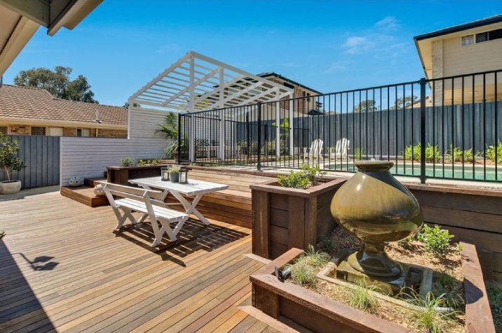 A large outdoor decking area with water fountain, table setting and pool behind.