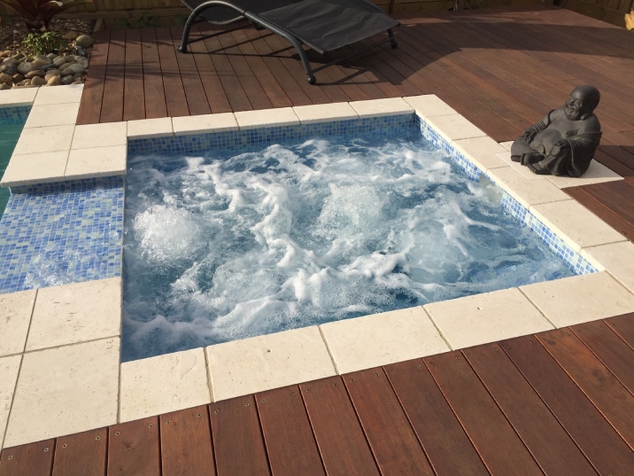 A square blue tiled spa that connects to a pool surrounded by decking.