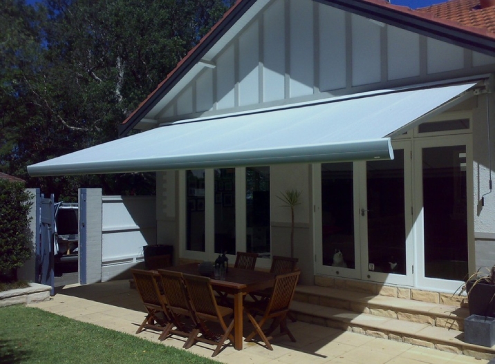 White house with roof tiles and sandstone pavers has a retractable shade sail coming off it.