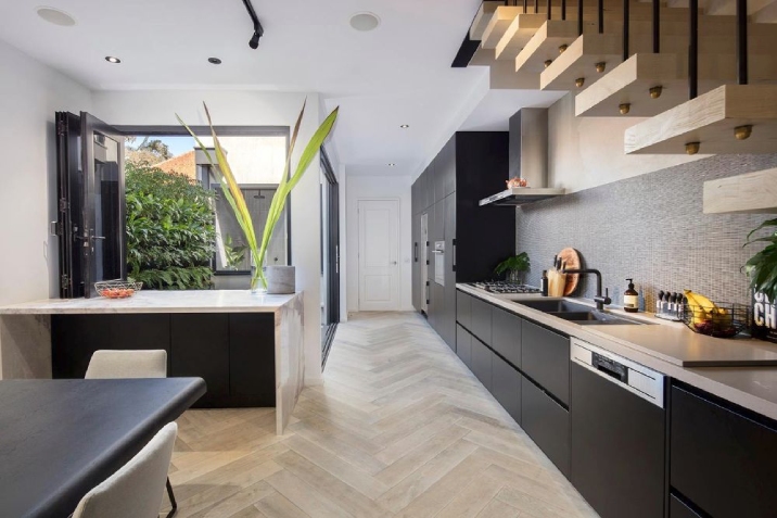 Long narrow kitchen with black cabinets, patterned timber floor and floating staircase.