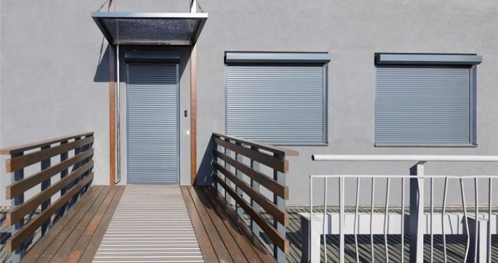 Timber walkway leading to a grey building with two windows and a door protected by roller shutters.
