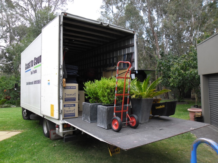 Mini truck with its back door open exposing several plants and a red trolley.