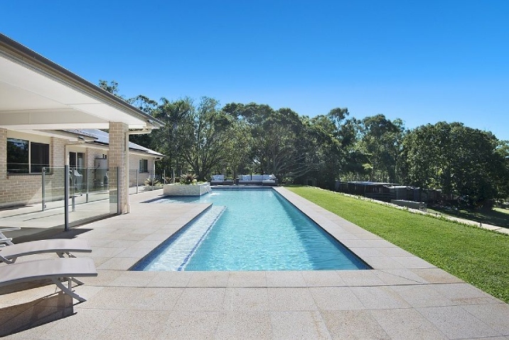 An L shaped blue pool with water feature, pergola on one side and grass on the other.